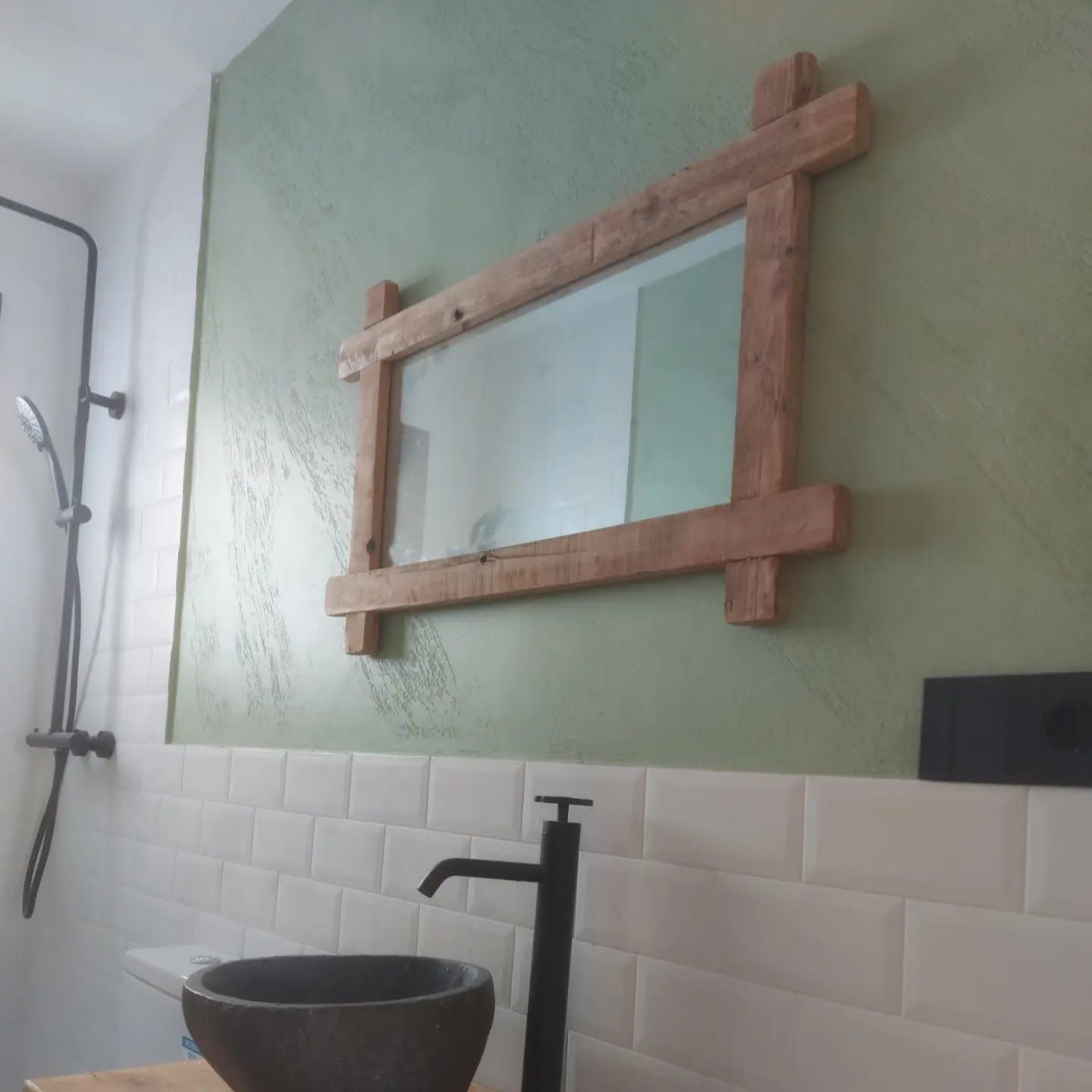 Baño con una pared verde, un espejo con marco de madera, azulejos blancos y un lavabo y grifo negros.