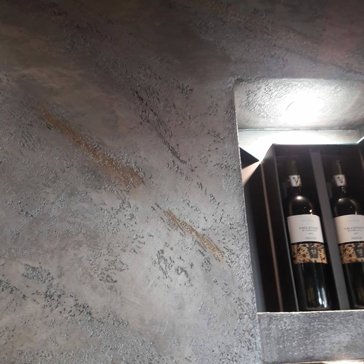 Dos botellas de vino en un estante con caja junto a una pared gris texturizada