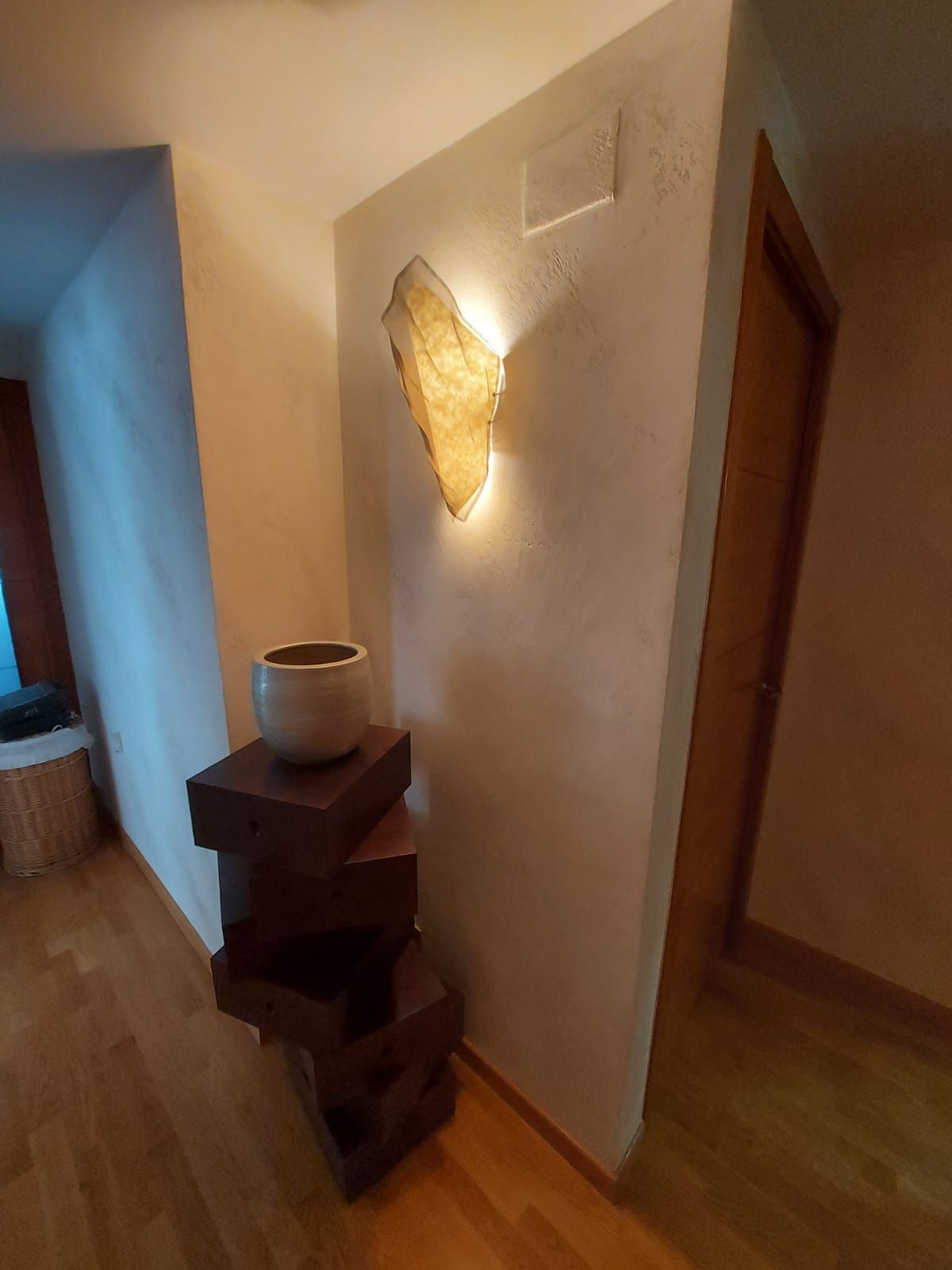 Pasillo con aplique de pared, suelo de madera y mesitas auxiliares de madera oscura apiladas, con una maceta.