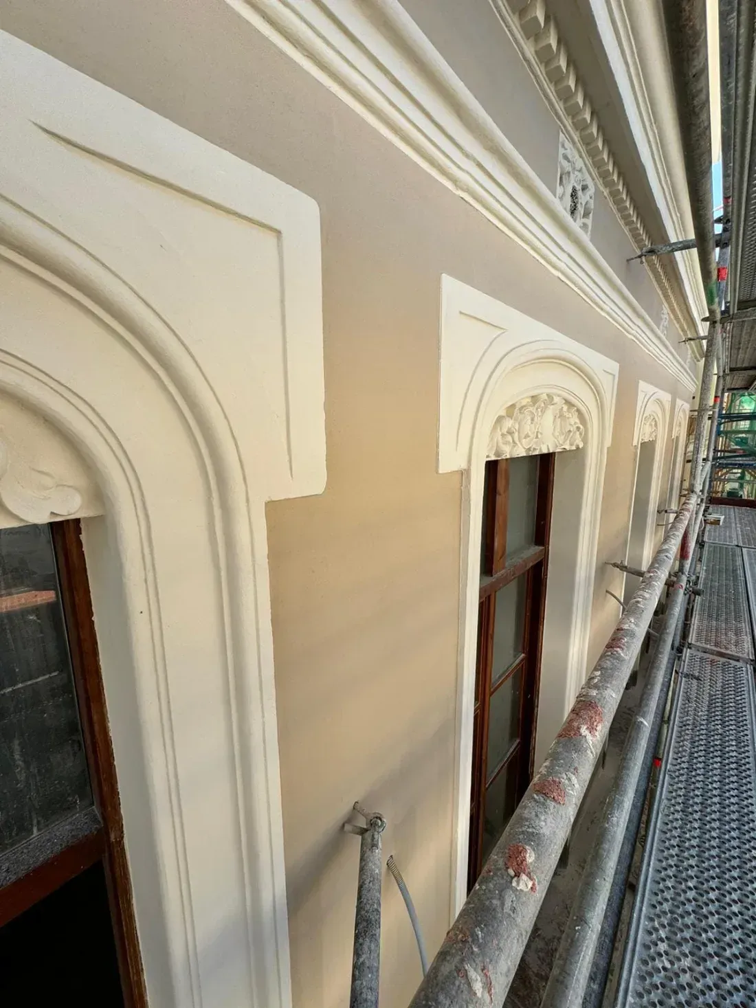 Pasarela de andamios junto a un edificio de color crema con ventanas arqueadas ornamentadas y molduras decorativas