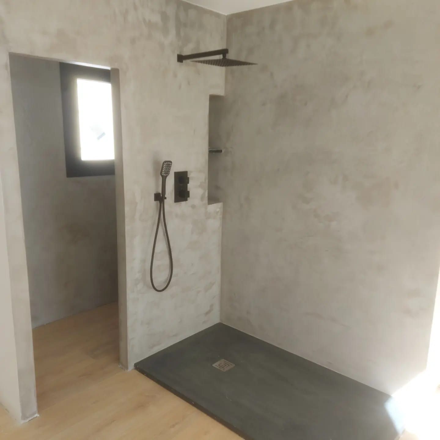Baño sin terminar con ducha, plato de ducha oscuro y una ventana pequeña en paredes grises.