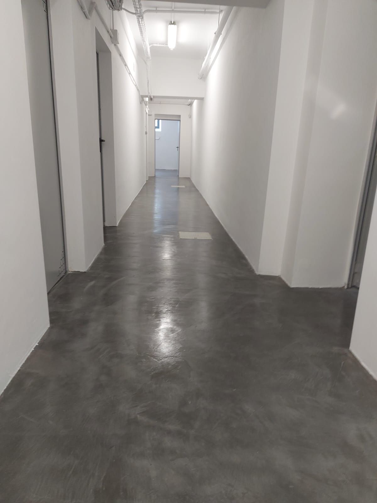 Pasillo blanco vacío con suelo gris y puertas a lo largo del lado izquierdo, iluminado por luces del techo