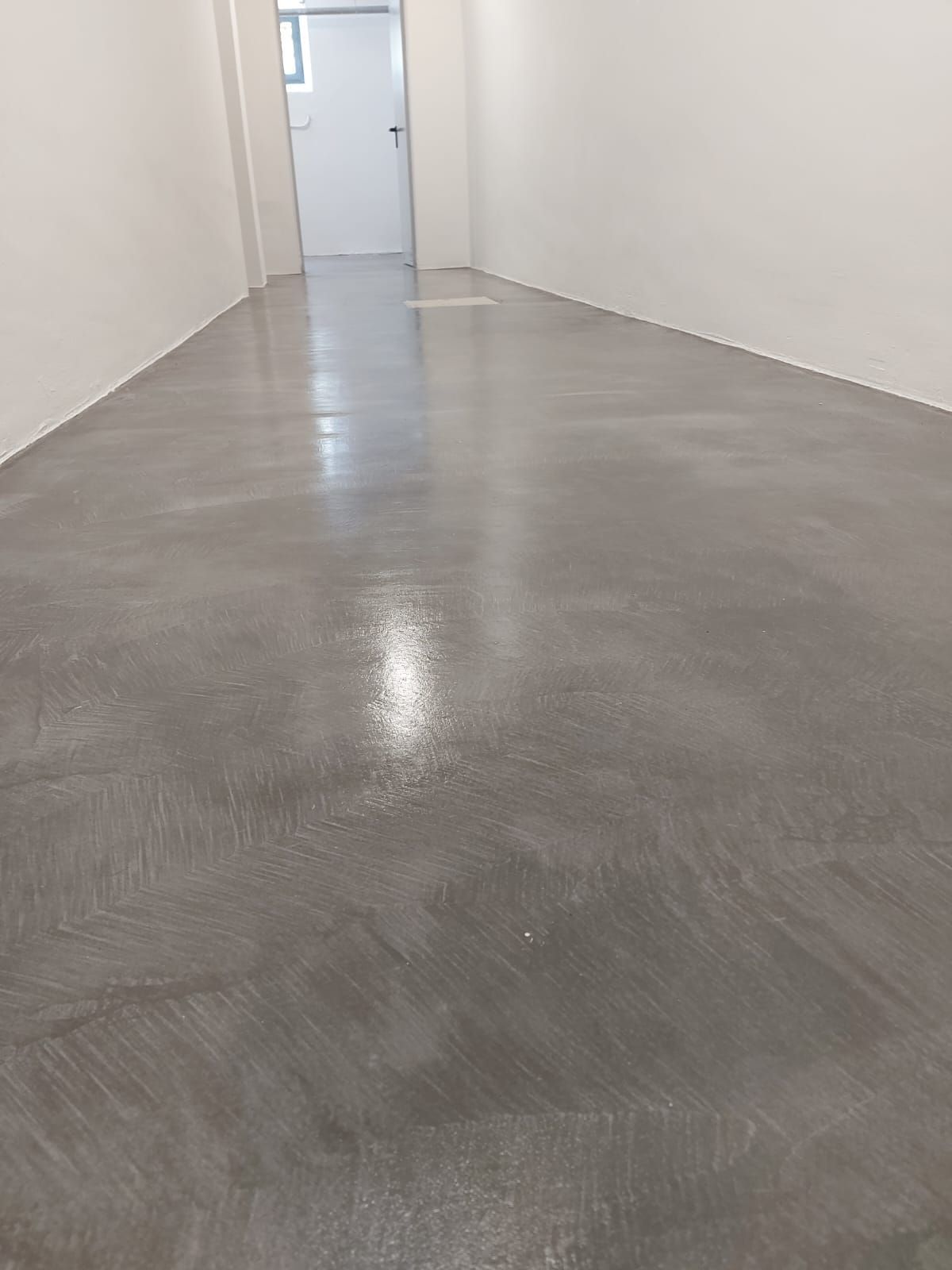 Pasillo vacío con suelo de hormigón gris pulido y paredes blancas que conduce a una puerta cerrada.
