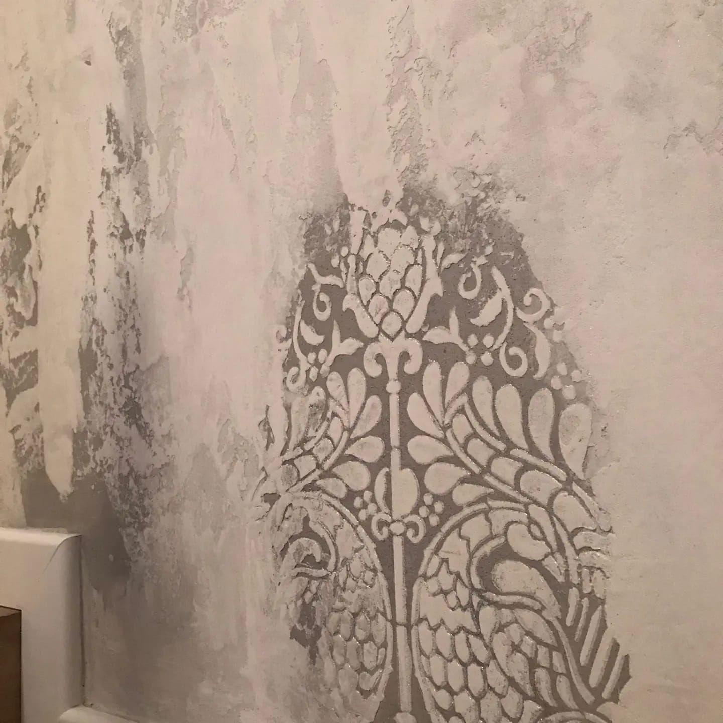 Pared de yeso dañada, con pintura descascarada y un elaborado patrón de plantilla gris