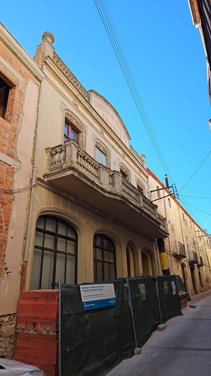 Calle estrecha con un histórico edificio beige, balcones y una valla verde bajo un cielo azul intenso