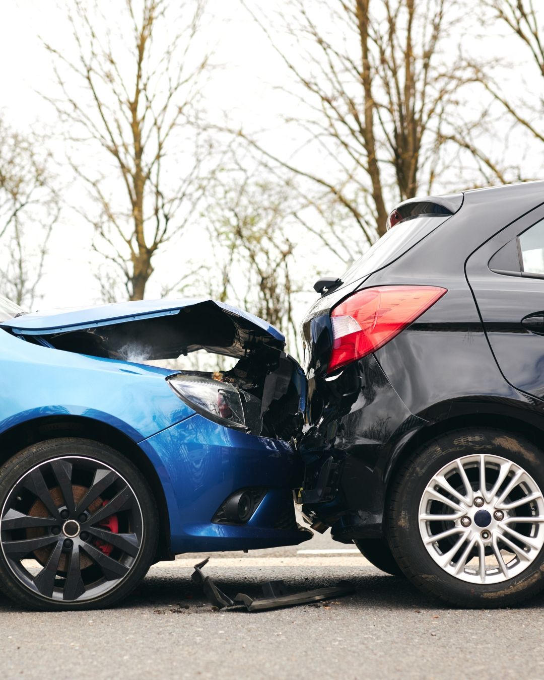 Un coche azul chocó contra la parte trasera de un coche negro en una carretera con árboles al fondo.