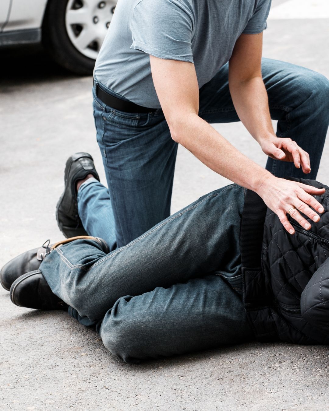 Una persona con una camiseta gris y vaqueros se arrodilla en el pavimento para ayudar a alguien que yace en el suelo.