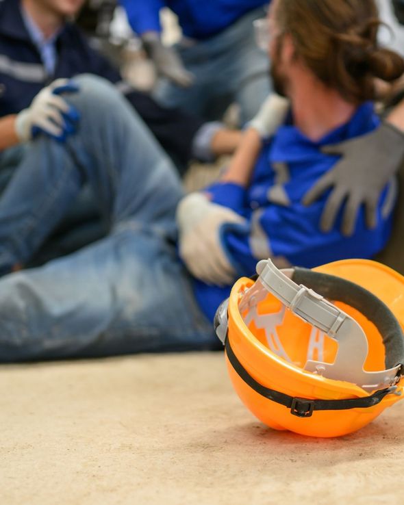 Un casco de seguridad de color naranja brillante se ve en primer plano mientras unos trabajadores con uniformes azules ayudan a un compañero herido en el suelo.