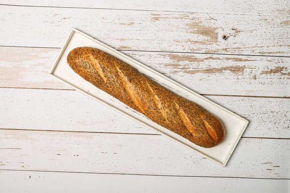 Ein Baguette mit Körnern auf einer weißen, rechteckigen Platte auf einem weißen Holztisch.