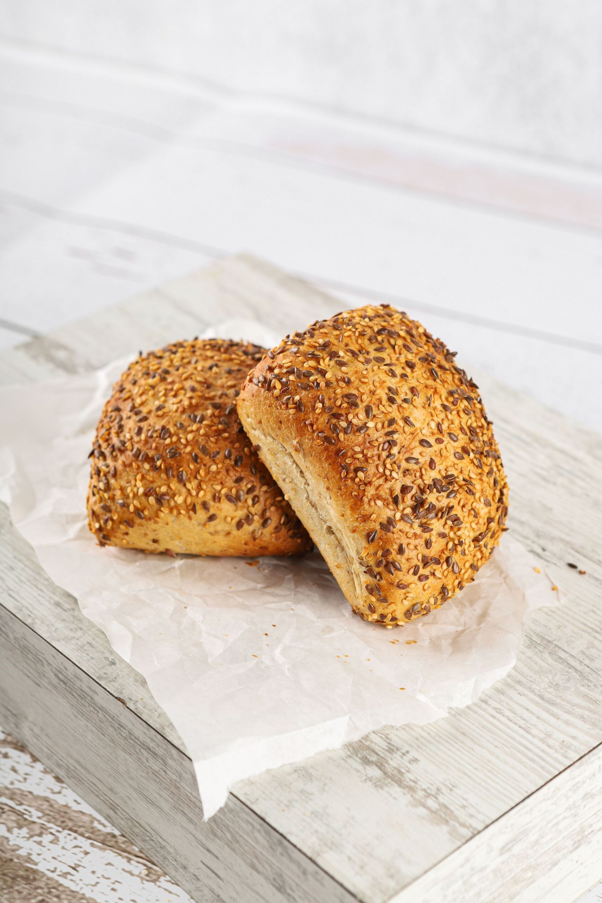 Zwei mit Körnern bestreute Brötchen auf Pergamentpapier, auf einer hellen Holzoberfläche platziert.