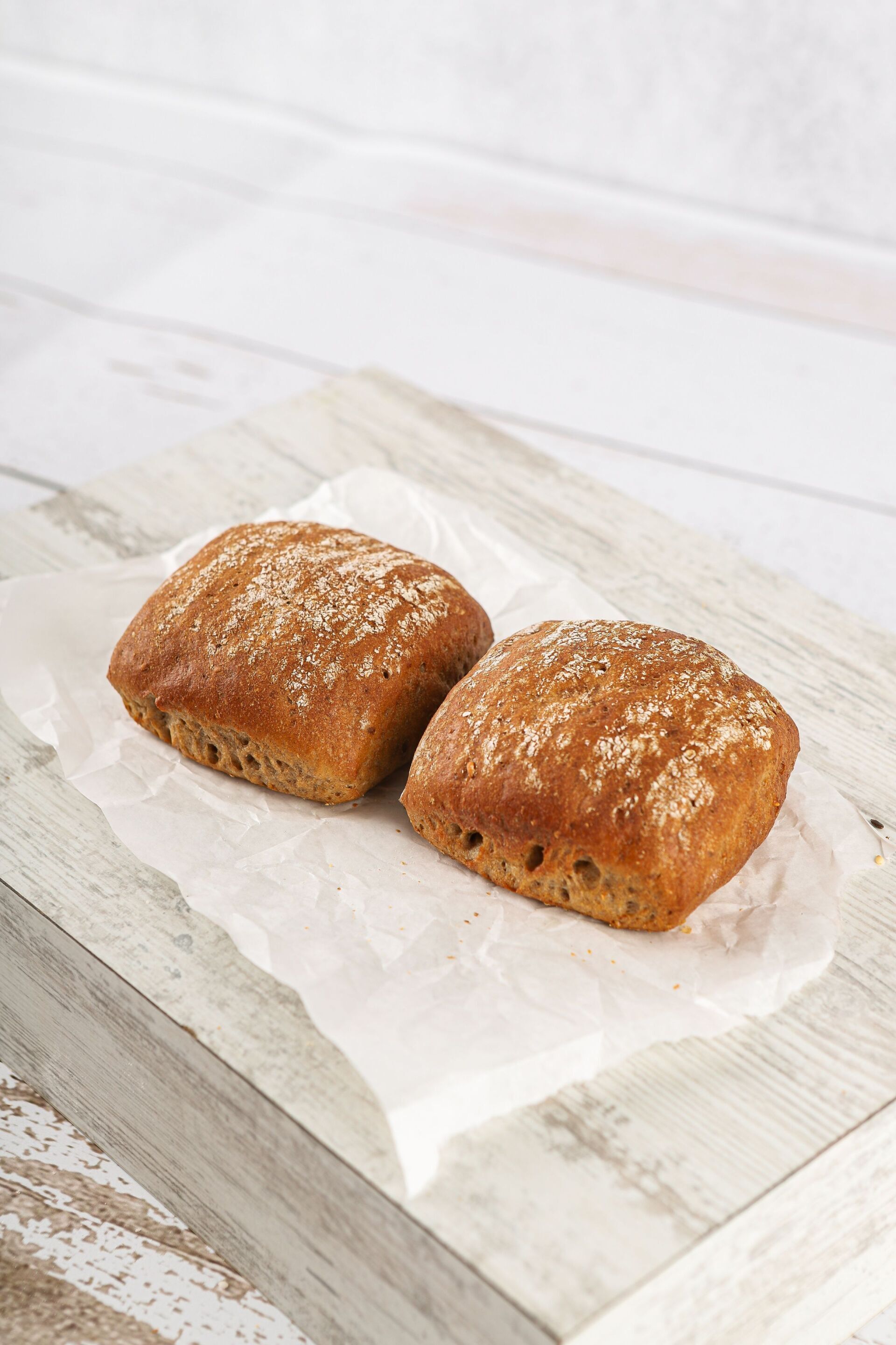 Zwei quadratische braune Brötchen auf weißem Pergamentpapier, auf einem Holzbrett.