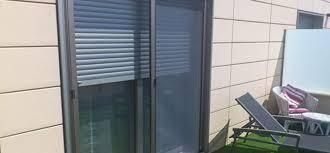 Puerta corredera de cristal con persianas cerradas, que da a una pequeña zona exterior con una tumbona y una mesa.