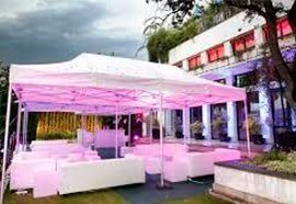 Montaje de fiesta al aire libre con toldo blanco, asientos e iluminación rosa cerca de un edificio.