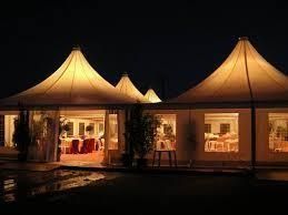 Tiendas de campaña iluminadas por la noche, con ventanas que dejan ver mesas y sillas en el interior, reflejándose en el suelo húmedo.