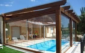 Piscina cubierta con paredes de cristal y estructura de madera, agua azul, día soleado.