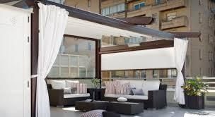 Terraza en la azotea con cortinas blancas, sofás, mesa de centro y toldo retráctil, frente a un edificio.