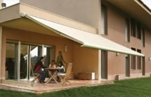 Un toldo da sombra a un patio donde hay gente sentada a una mesa. Exterior del edificio, césped verde.