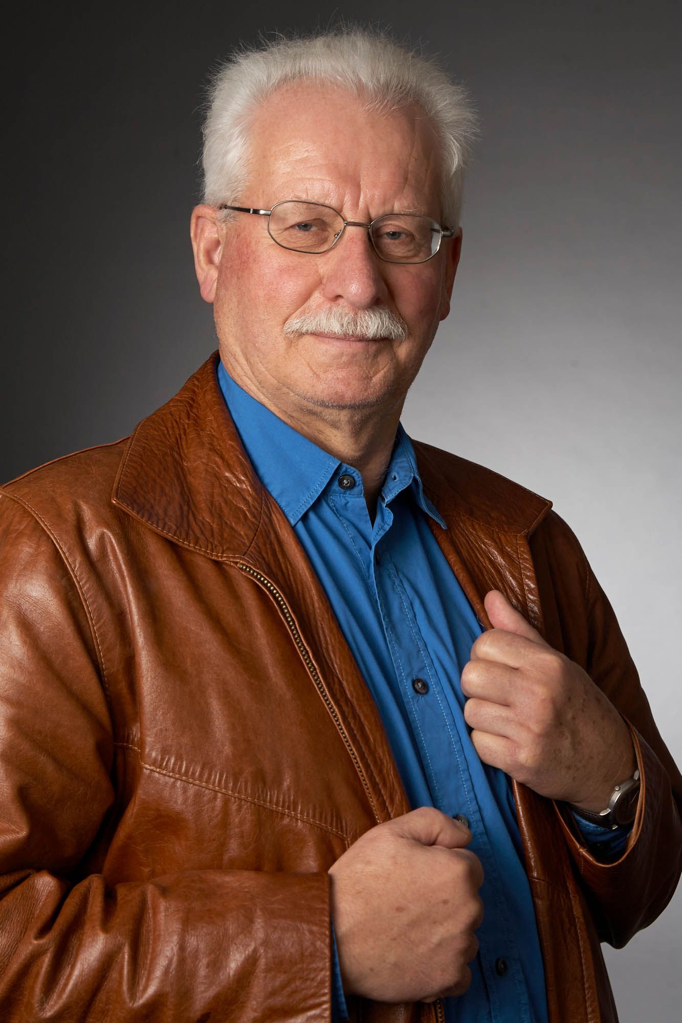 Ein Mann mit Brille und Schnurrbart trägt eine braune Lederjacke und ein blaues Hemd.