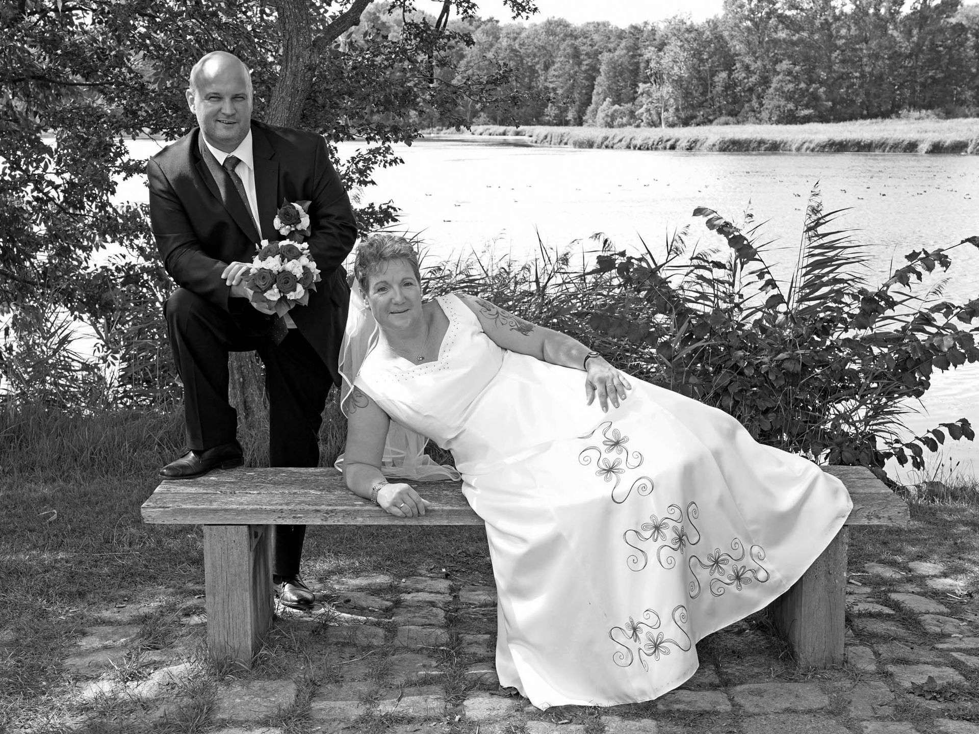 Eine Braut und ein Bräutigam posieren auf einer Bank neben einem See für ein Foto.