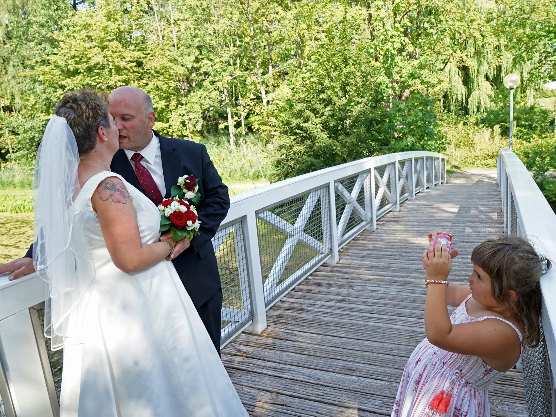 Ein kleines Mädchen macht ein Foto von Braut und Bräutigam auf einer Brücke.