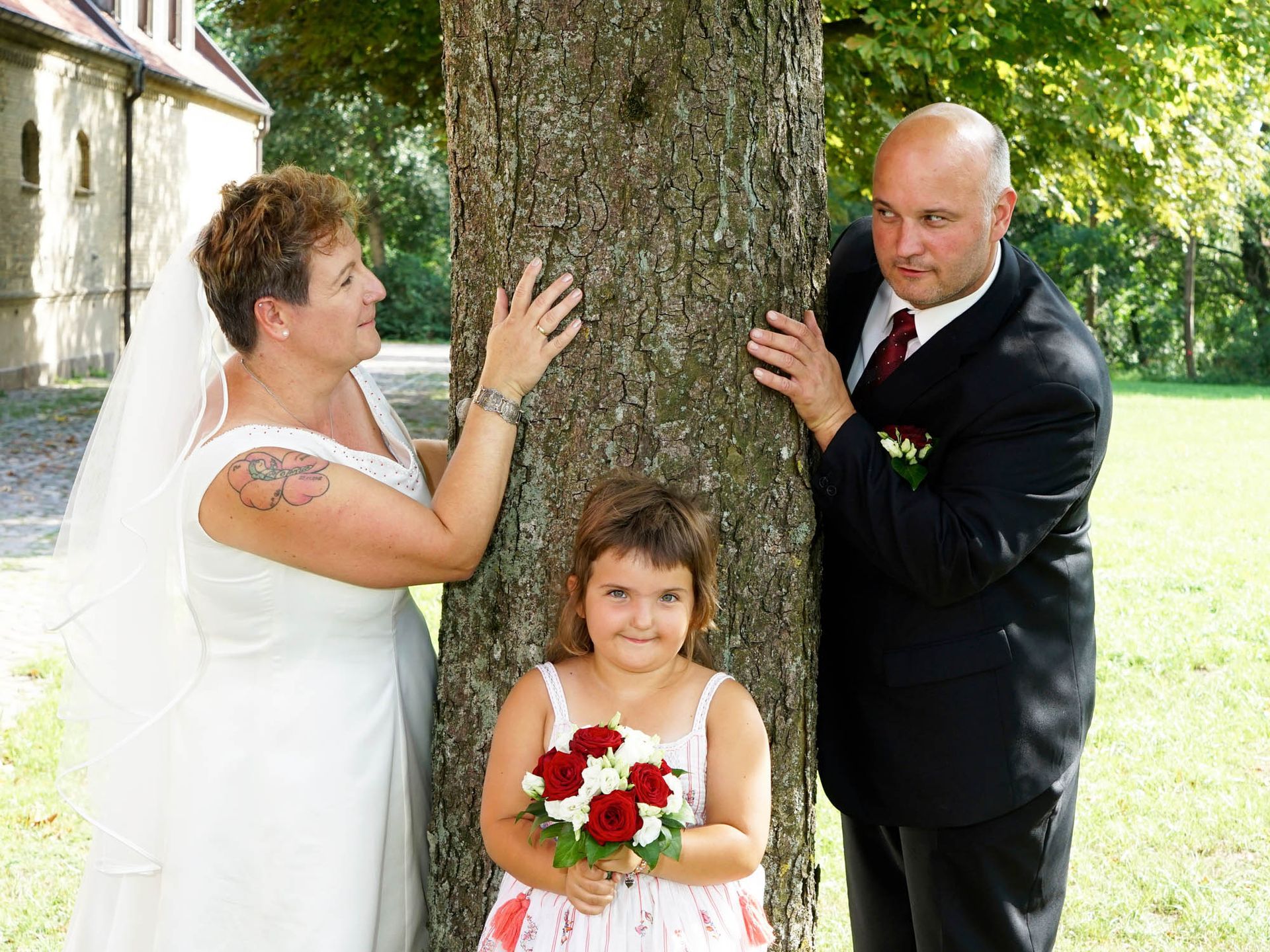 Eine Braut und ein Bräutigam stehen neben einem Baum, ein kleines Mädchen hält einen Blumenstrauß.