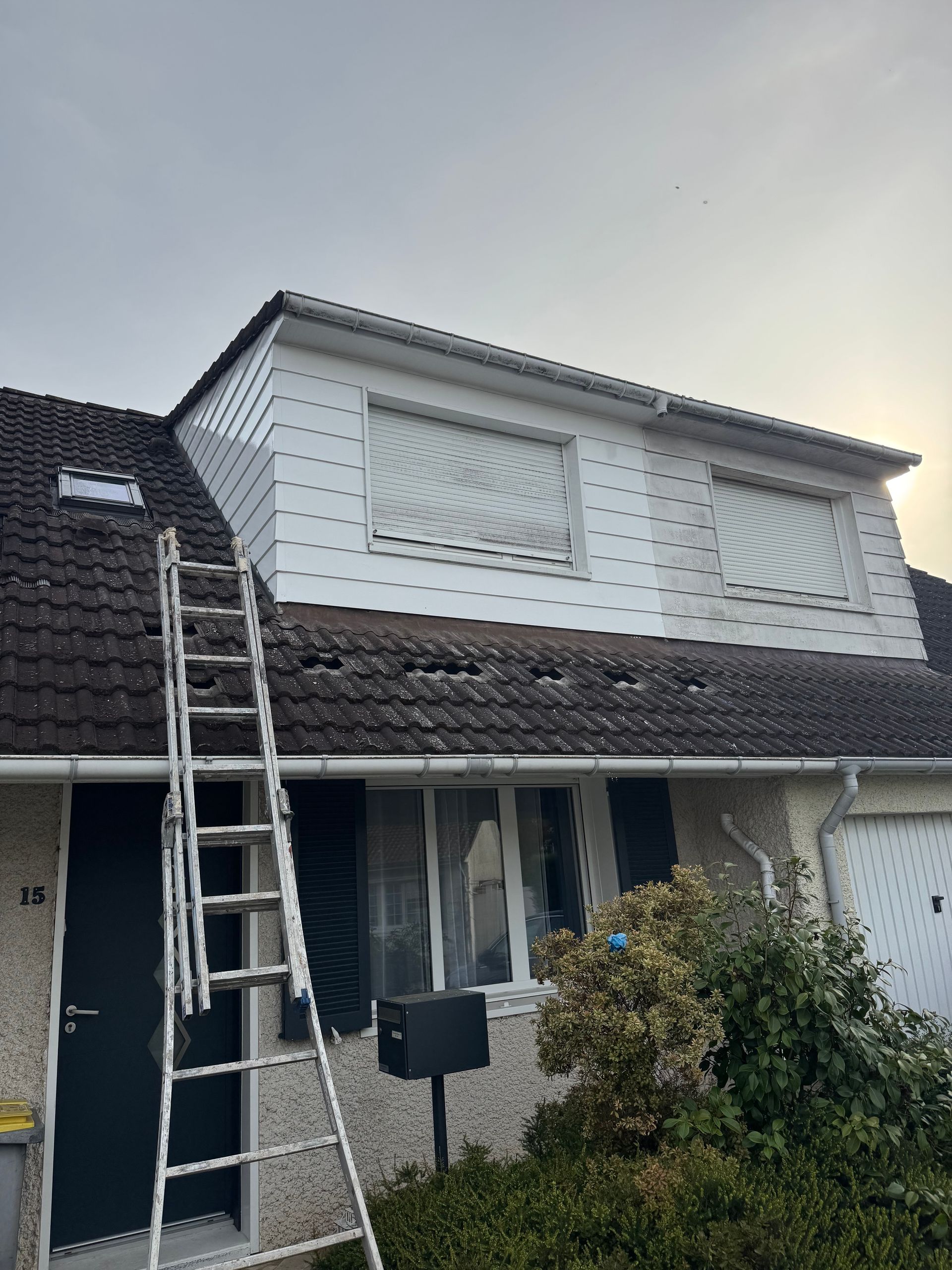 Une échelle appuyée contre le toit de tuiles sombres d'une maison à deux étages avec un bardage blanc et des volets roulants.
