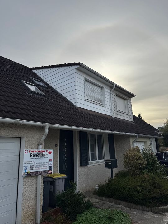 Une échelle appuyée contre le toit de tuiles sombres d'une maison à deux étages avec un bardage blanc et des volets roulants.