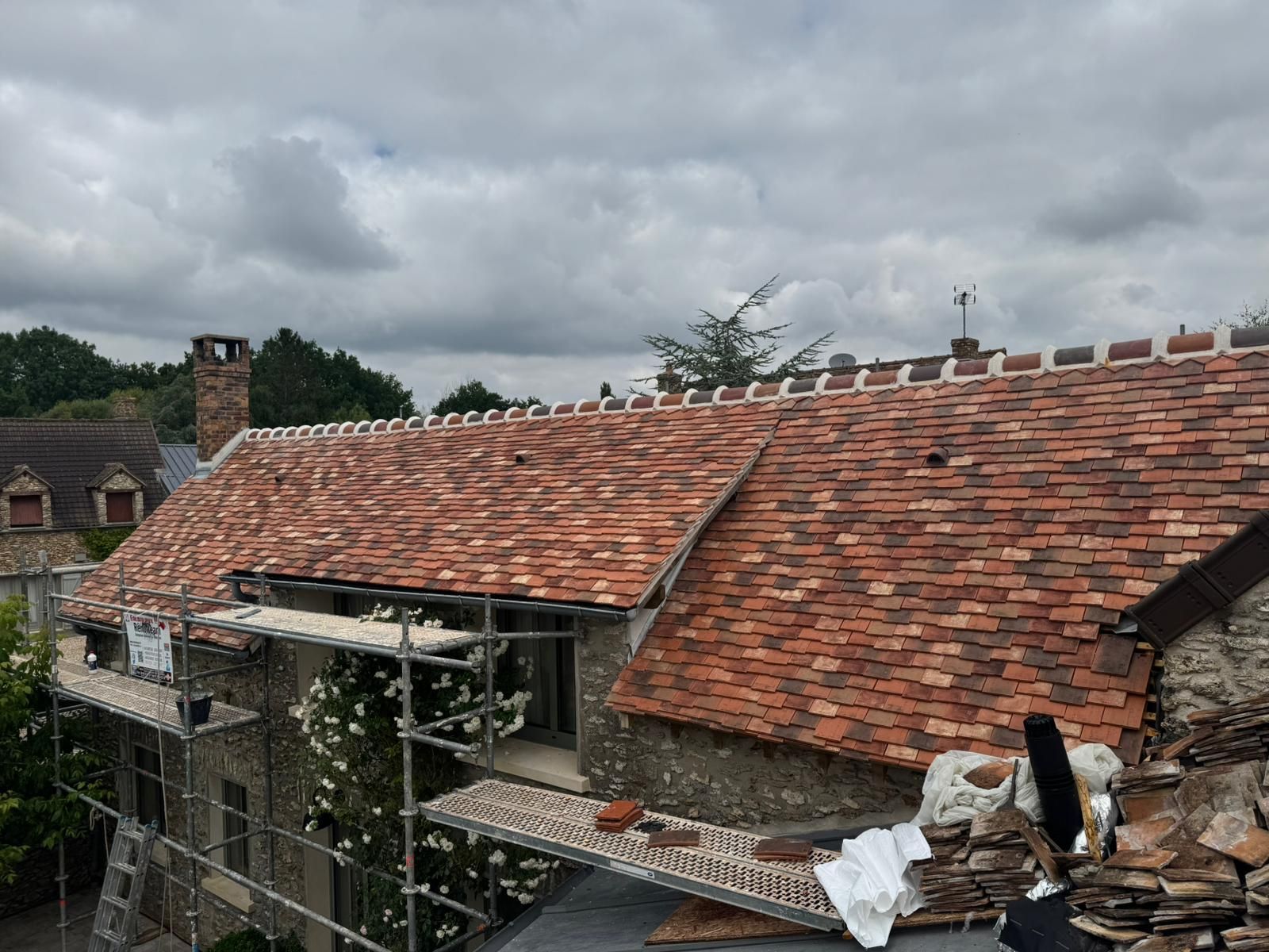 Des échafaudages contre un bâtiment en pierre au toit de tuiles multicolores fraîchement posé, sous un ciel nuageux.