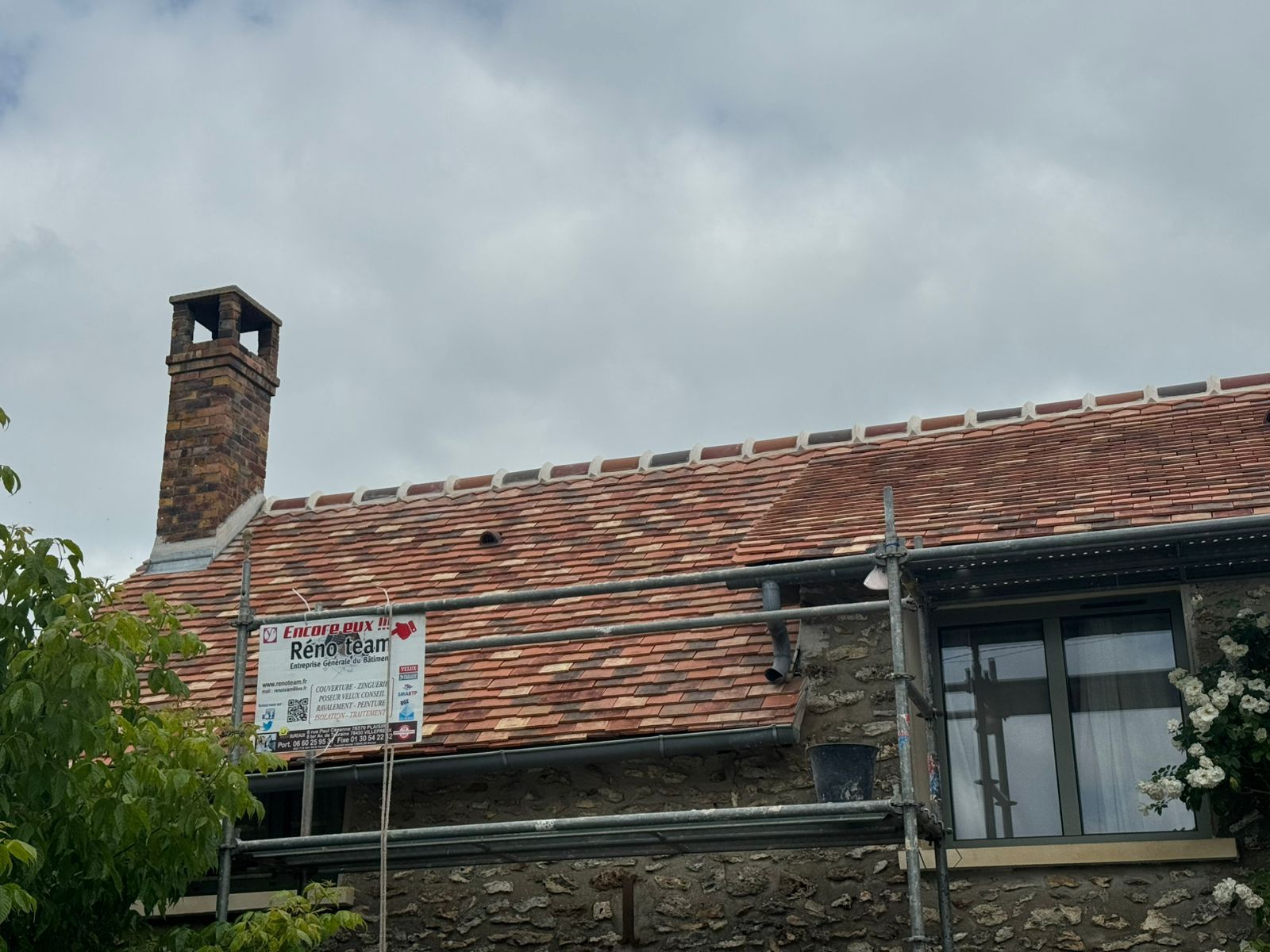 Vieille maison en pierre avec toit de tuiles rouges, cheminée et petite pancarte sous un ciel nuageux