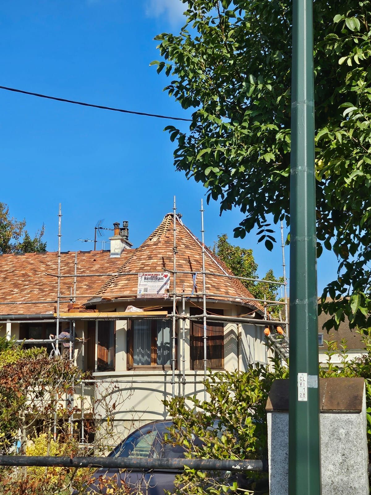 Maison au toit de tuiles rouges en cours de réparation, partiellement cachée par un arbre et un poteau.