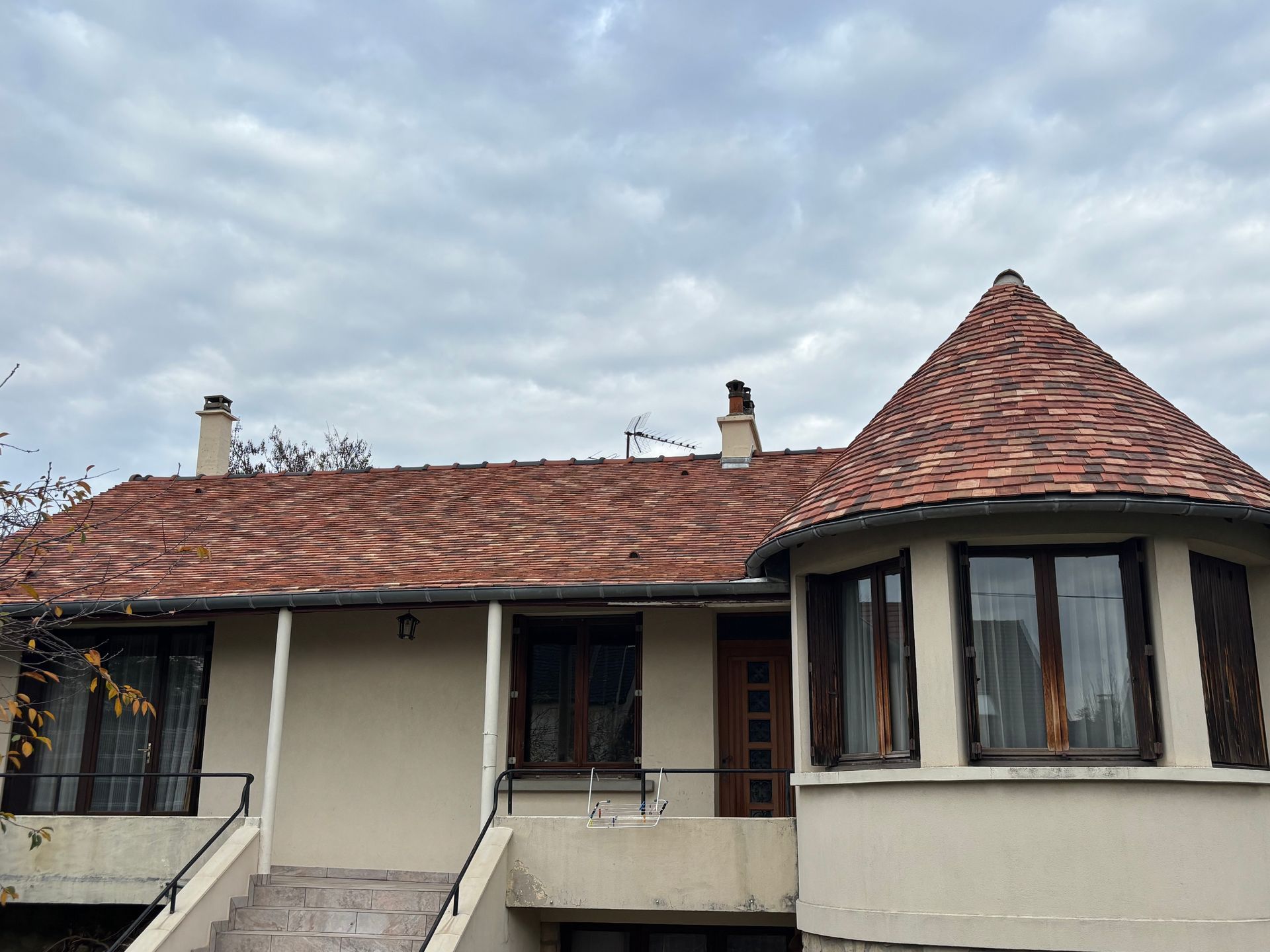 Une maison avec un toit de tuiles en terre cuite texturée, comprenant une tourelle sur la droite, sous un ciel nuageux.