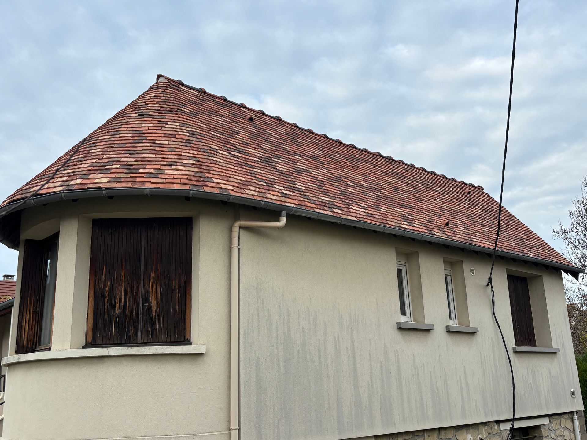 Petite maison beige au toit de tuiles rouges et aux volets sombres sous un ciel nuageux