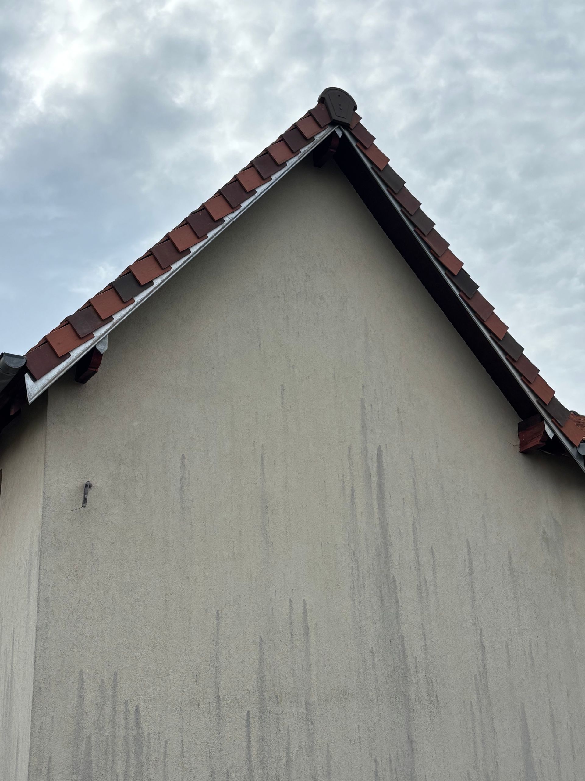 Gros plan sur le pignon d'une maison aux murs en stuc beige et au toit de tuiles rouges et noires sous un ciel nuageux