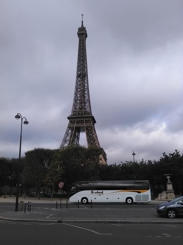 La tour Eiffel se dresse derrière une rangée d'arbres et un bus touristique blanc garé dans une rue de la ville sous un ciel nuageux.