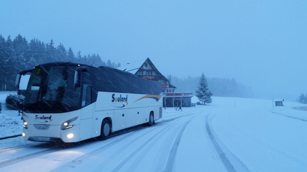 Un autocar blanc de la marque Sculand est stationné sur une route enneigée, avec un bâtiment et une forêt en arrière-plan, par une journée d'hiver.