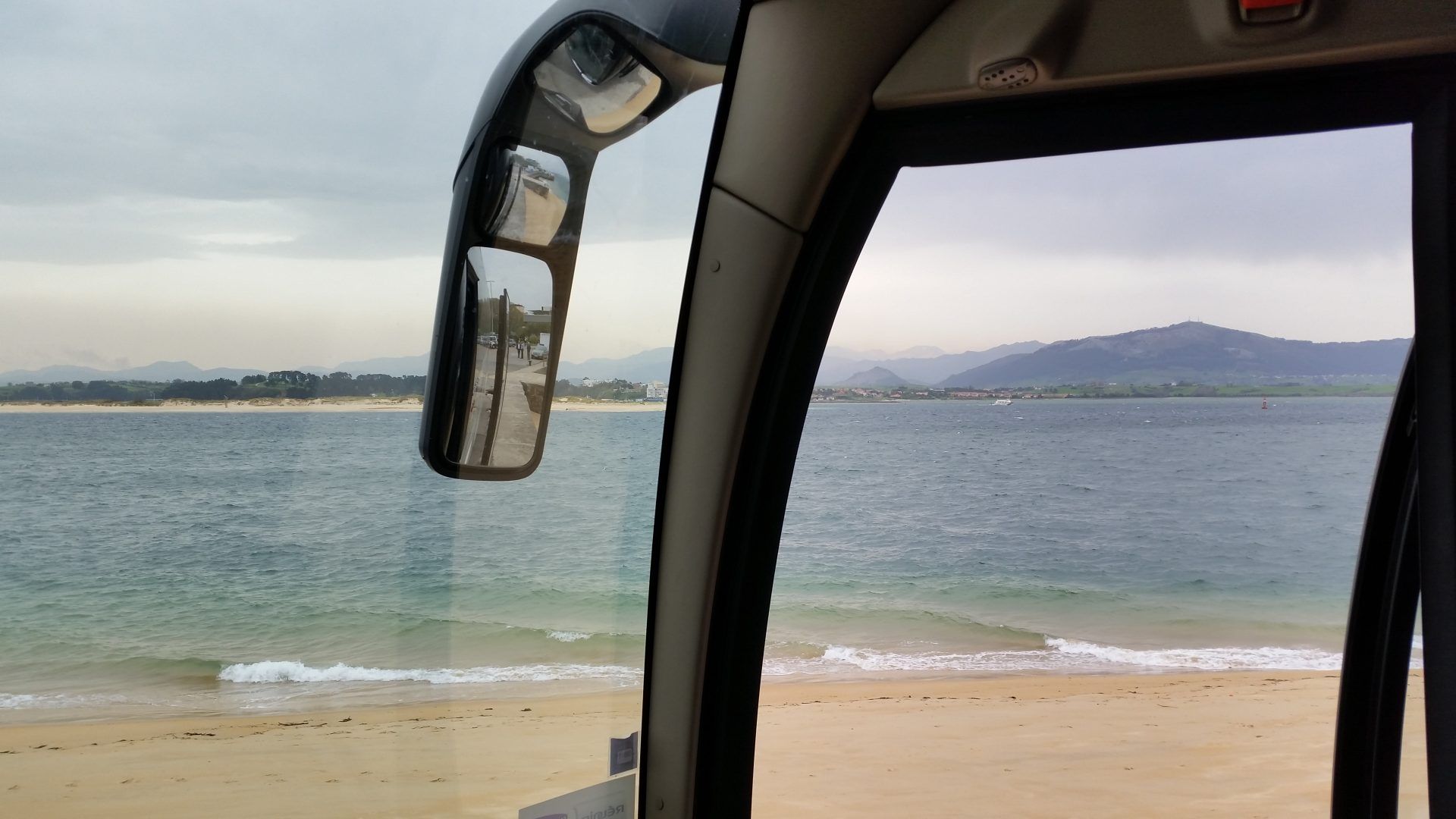 Vue par la fenêtre d'un bus montrant une plage de sable fin, une eau calme et des montagnes au loin sous un ciel nuageux.