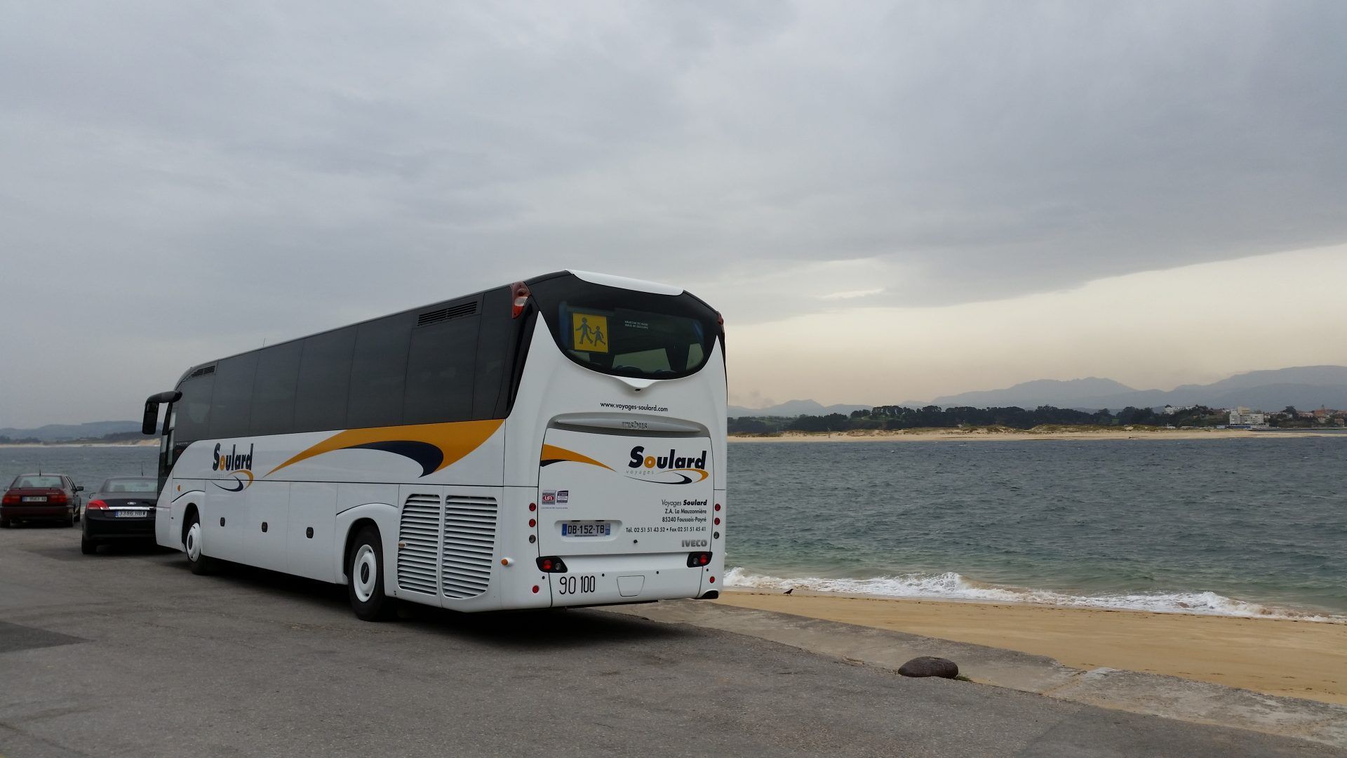 Un bus touristique blanc avec des touches de jaune est garé sur une route longeant une plage de sable fin et des eaux calmes sous un ciel nuageux.