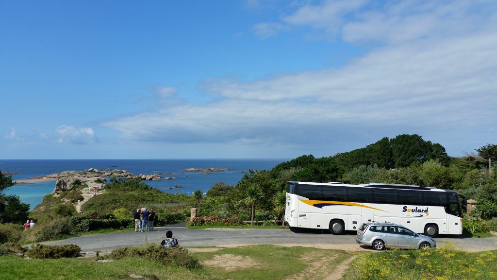 Un bus touristique blanc et une voiture argentée sont garés sur un promontoire herbeux surplombant une baie côtière aux eaux bleues et aux rivages rocheux.