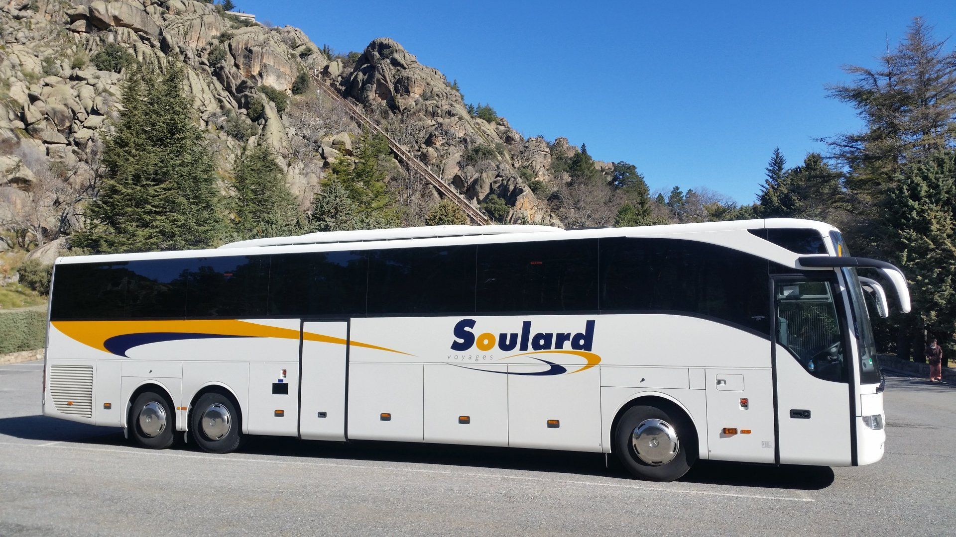 Un bus touristique Soulard blanc, orné de touches de jaune et de bleu, est stationné dans un paysage montagneux sous un ciel dégagé.
