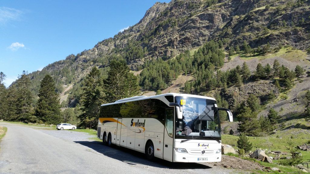 Un bus touristique blanc garé en bord de route, devant un grand versant montagneux boisé, par une journée ensoleillée.