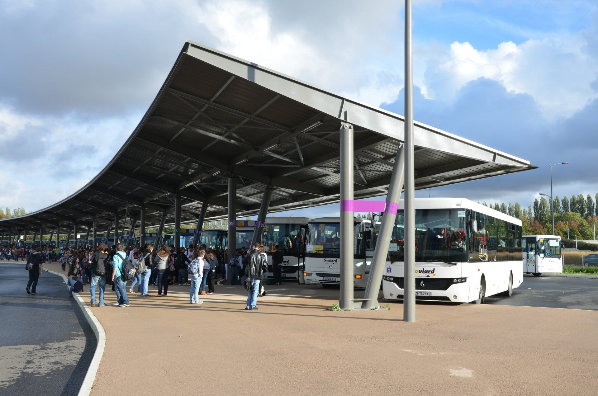 Une gare routière moderne avec un grand auvent incurvé, de nombreux bus blancs et des personnes debout à proximité sous un ciel nuageux.