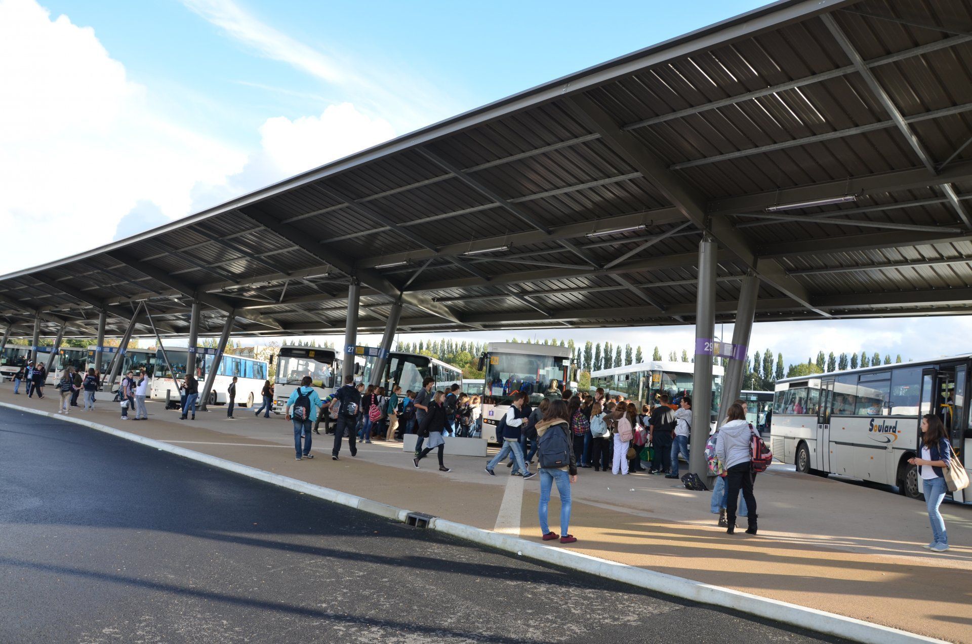 Des personnes patientent sous un grand auvent moderne à une gare routière extérieure où plusieurs autocars sont stationnés.