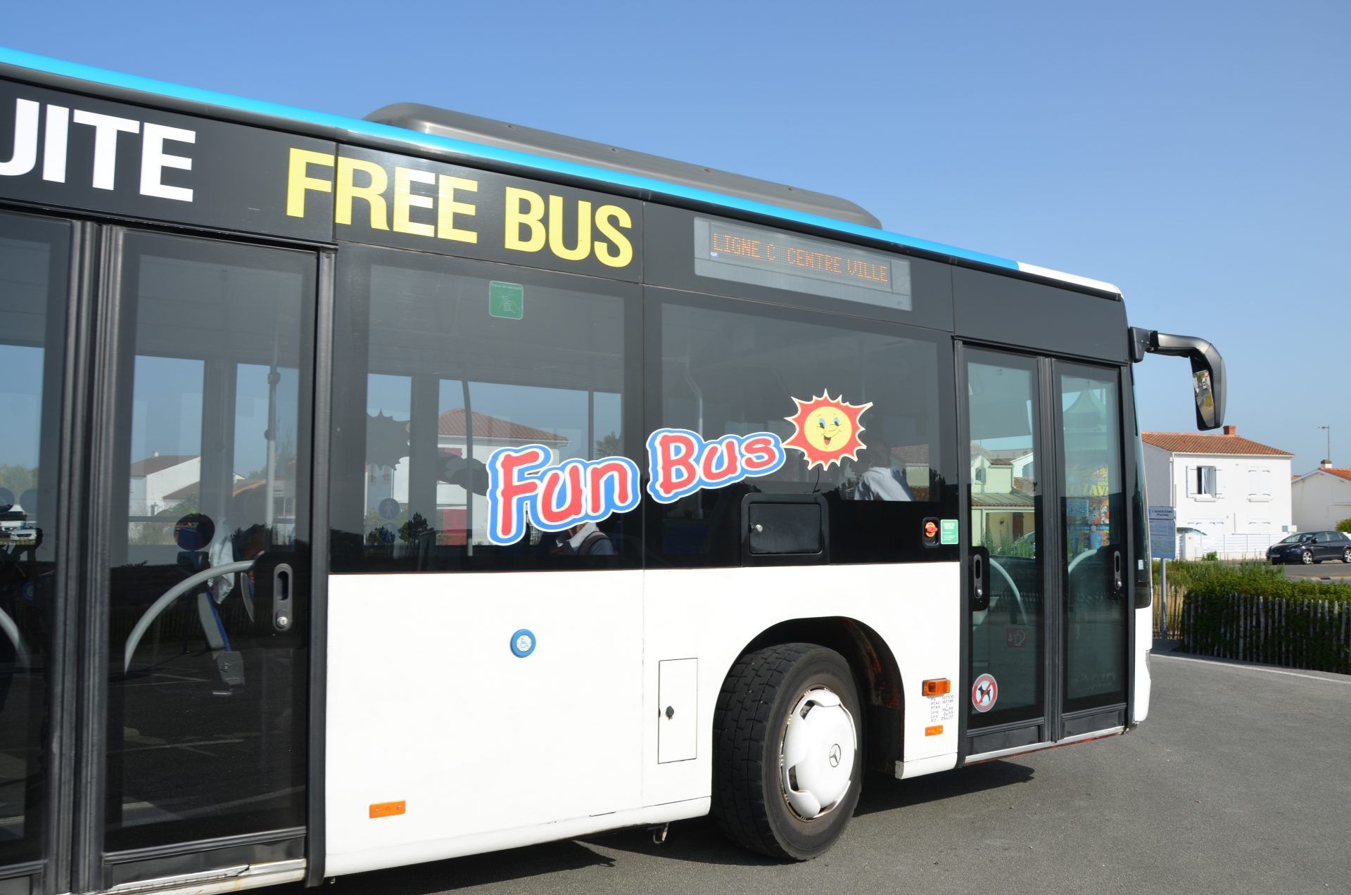 Un bus blanc portant l'inscription « FREE BUS » et « Fun Bus » ainsi qu'un symbole de soleil sur le côté, est stationné à l'extérieur par une journée ensoleillée.