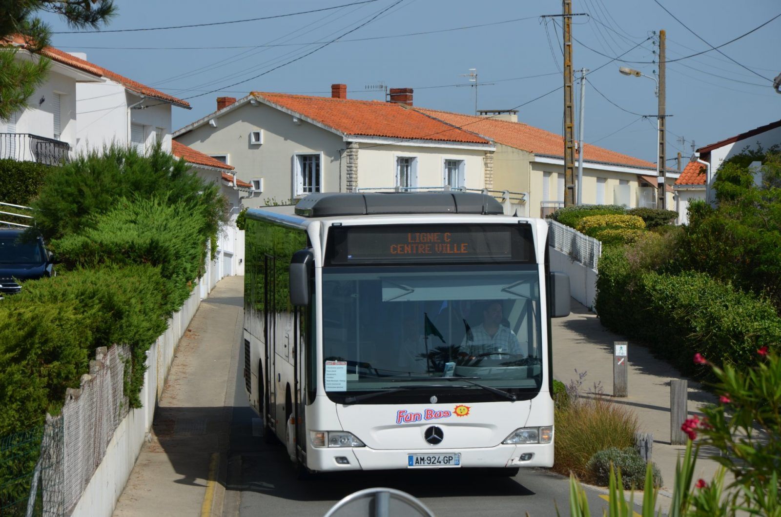 Un autobus Mercedes blanc circule dans une rue résidentielle étroite bordée de maisons et de verdure.