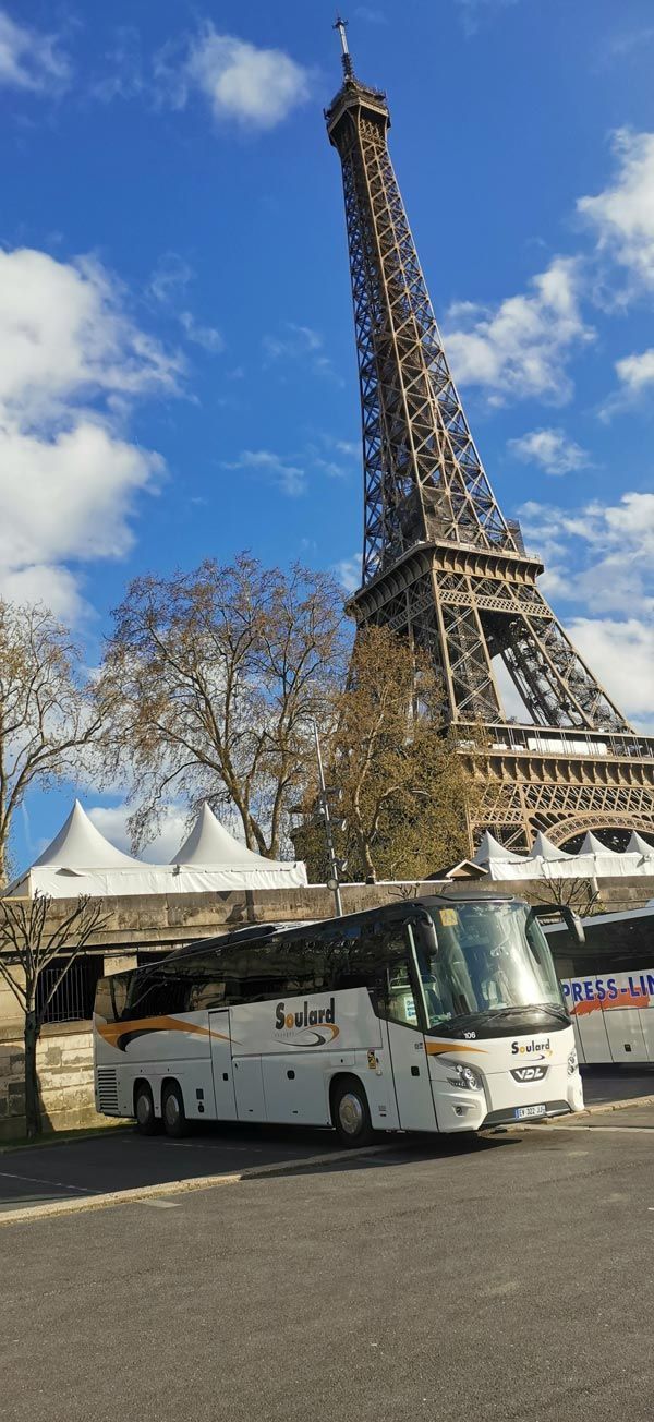 Un bus touristique garé devant la Tour Eiffel à Paris, sous un ciel bleu parsemé de nuages ​​épars.