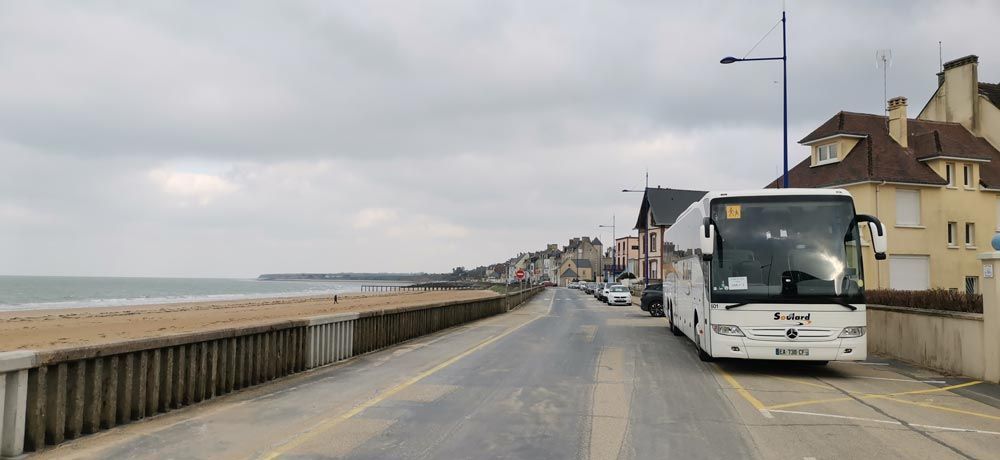 Un bus blanc garé sur une route côtière, à côté d'une plage de sable et de maisons, sous un ciel nuageux.