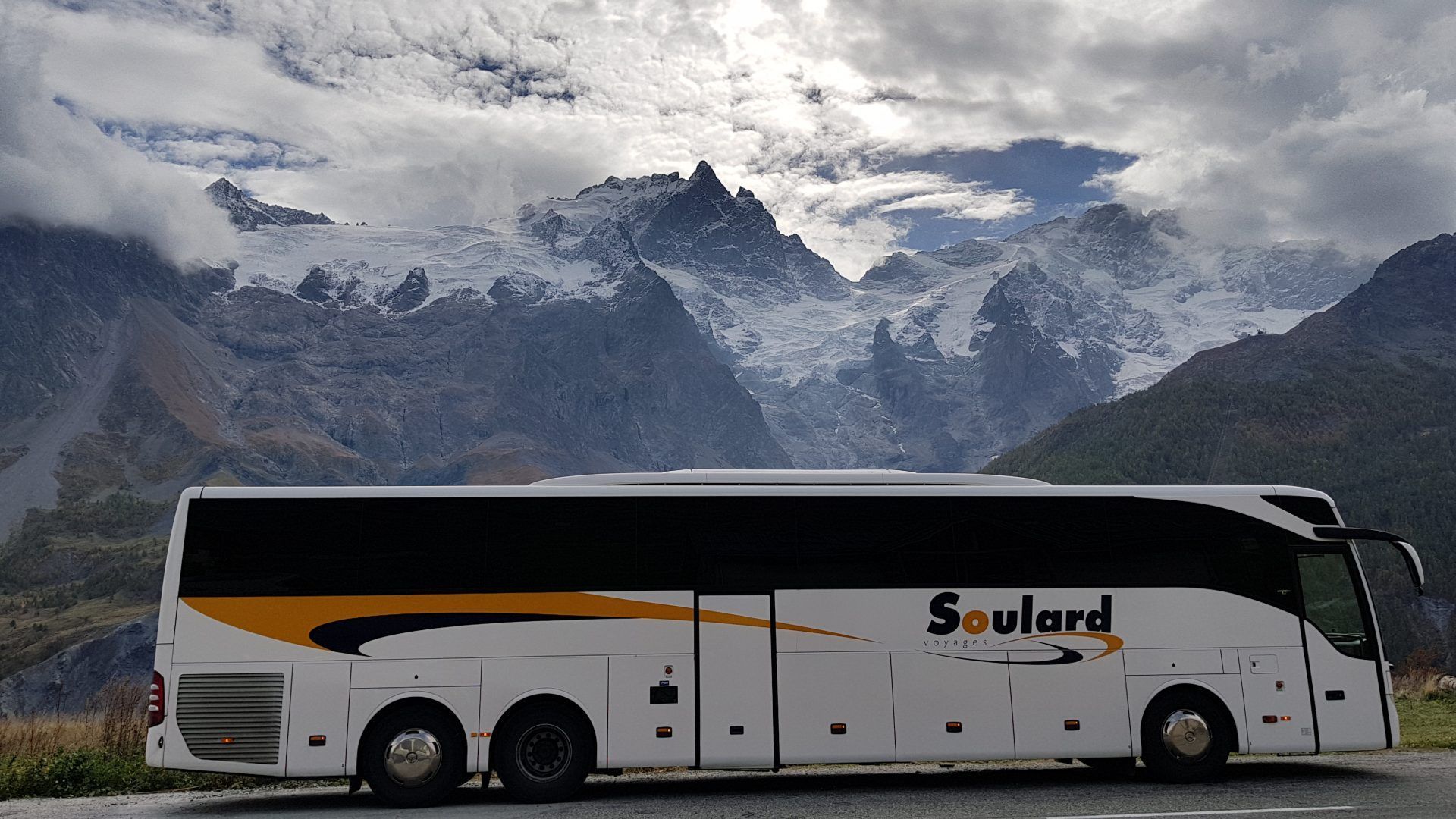 Un bus touristique Soulard blanc et orange, garé devant un décor de montagnes enneigées sous un ciel nuageux.