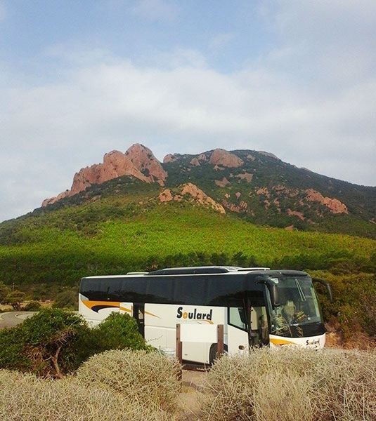 Un bus touristique blanc avec des touches de jaune est garé devant une montagne verdoyante aux formations rocheuses rouges apparentes.