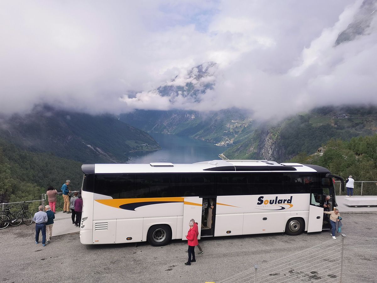 Un bus touristique blanc portant l'inscription « Soulard » est stationné à un point de vue panoramique avec des montagnes et un fjord en arrière-plan.