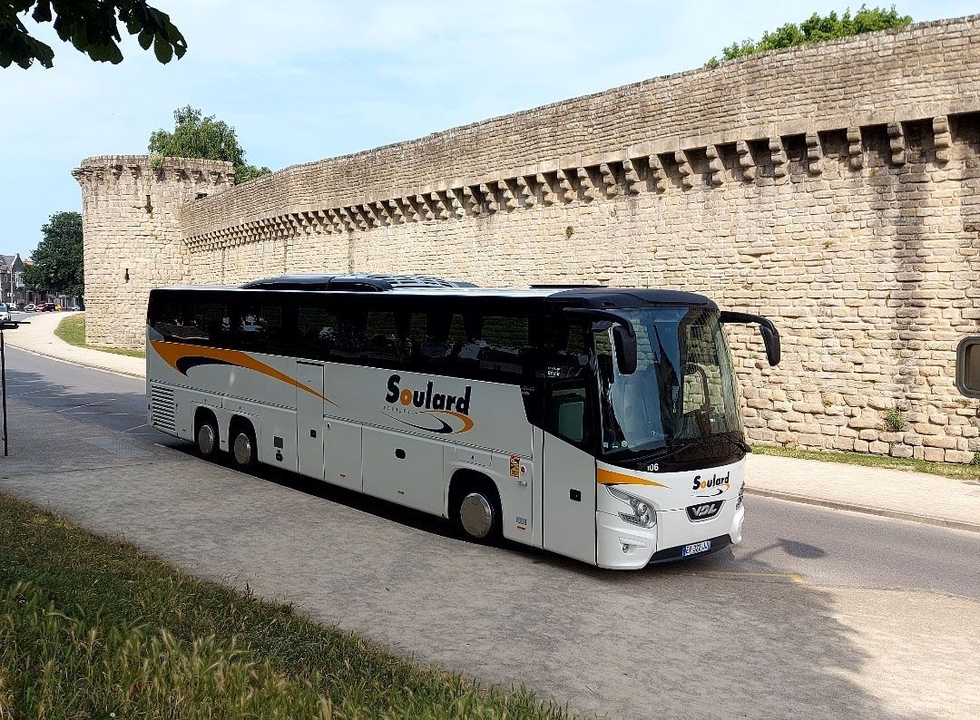 Un bus touristique blanc et noir avec des touches de jaune circule sur une route longeant un haut mur défensif en pierre historique.